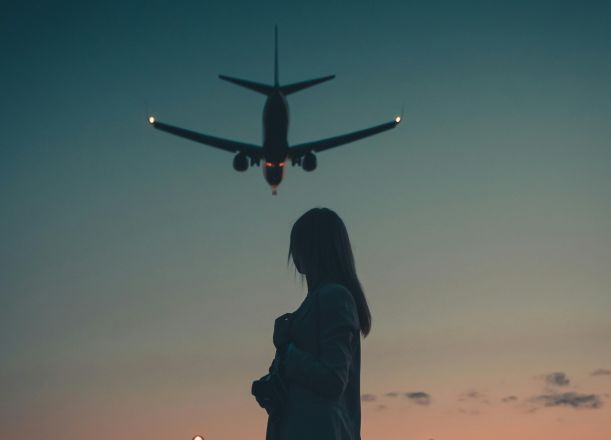 A silhouette of a woman standing as an airplane flies overhead during dusk near an airport.